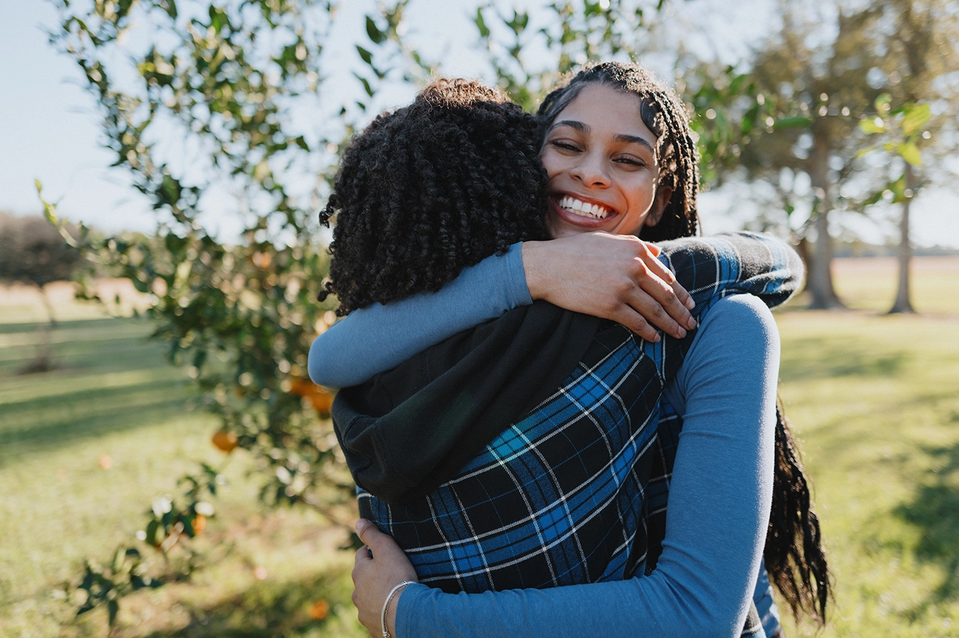 A woman smiling and giving her friend a warm hug surrounded by orange trees.