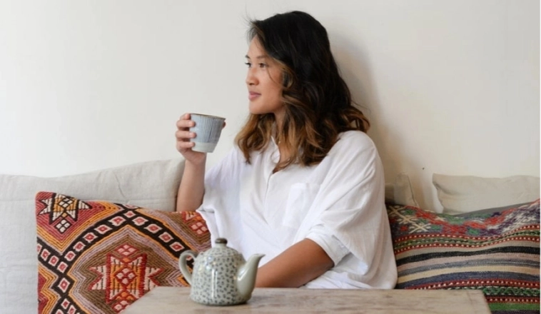 Woman with dark hair drinking a cup of tea and sitting on the couch surrounded by patterned pillows.