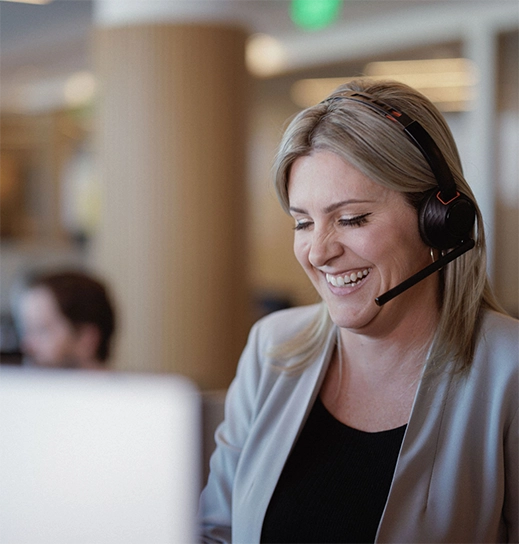 A white woman with blonde hair and a beige jacket sits in front of a laptop wearing a headset, sharing a laugh with a client on the phone.