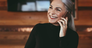 A mature woman in a black shirt with long silver hair smiles while talking on her phone.