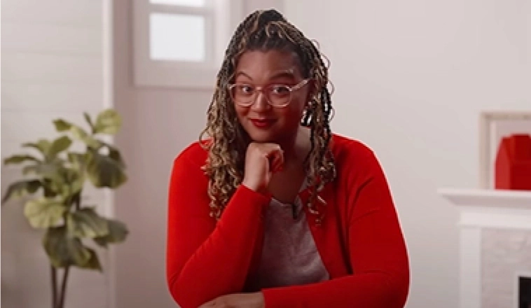 The Red Desk spokeswoman sitting at a red desk wearing a a red sweater with her chin resting on her hand.