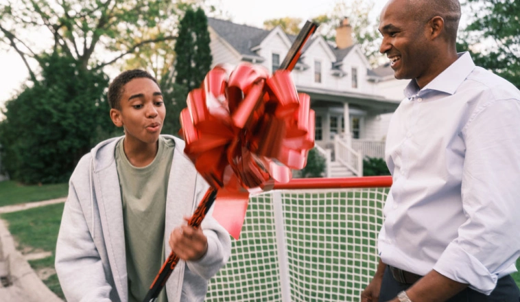 A teenage boy holds a gift of sports equipment with a big red bow on it in a backyard while his dad looks on.