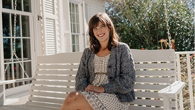 A white woman with long dark hair wearing a print dress relaxes on a white wooden porch swing.