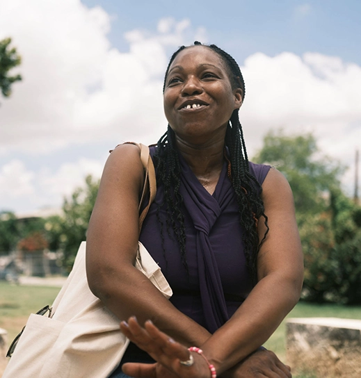 A Black woman with long braided hair and a tote bag over her shoulder looks engaged in conversation outdoors.