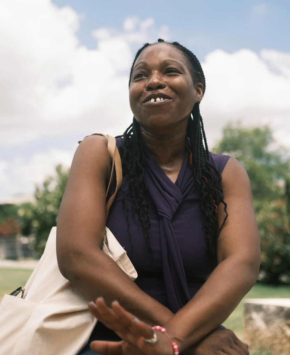 A Black woman with long braided hair and a tote bag over her shoulder looks engaged in conversation outdoors.