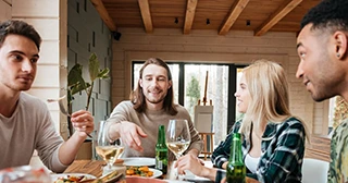 Four friends enjoy wine and a meal together around a table in a kitchen with wood paneling and ceiling.