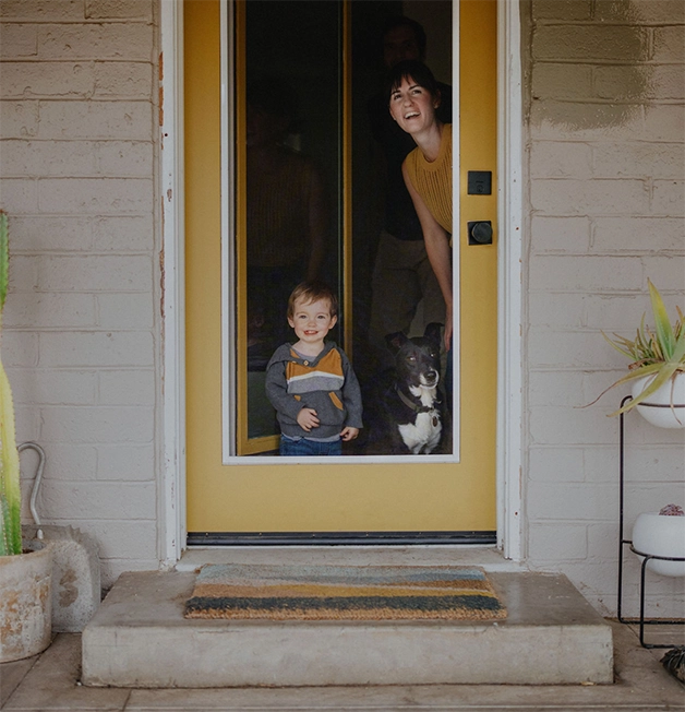 A smiling young white woman with short dark hair looks out the front door of her home along with her toddler son and black and white dog.