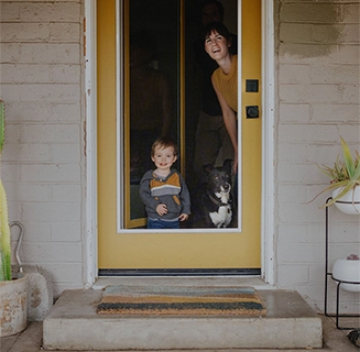 A smiling young white woman with short dark hair looks out the front door of her home along with her toddler son and black and white dog.