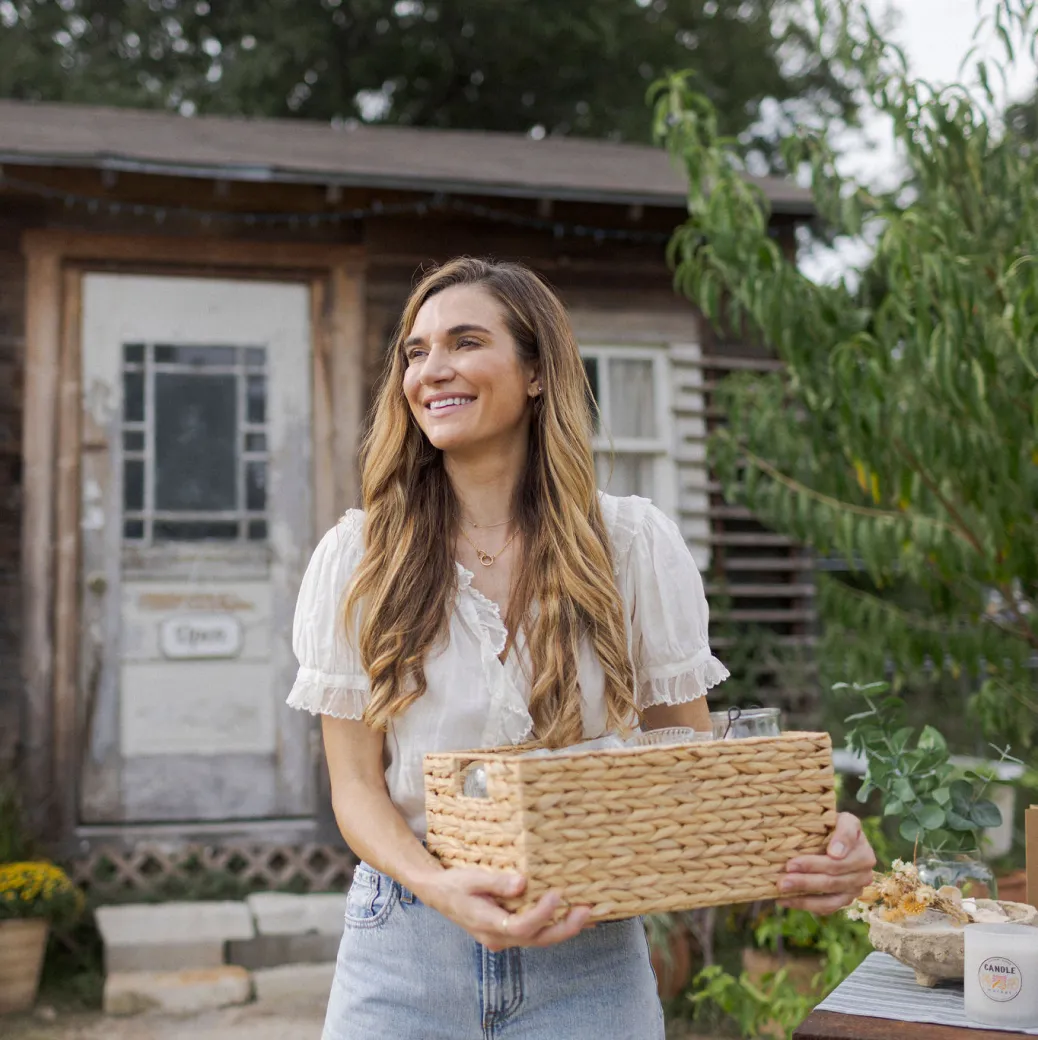 A white woman stands in front of her small business, holding a basket of handmade goods.