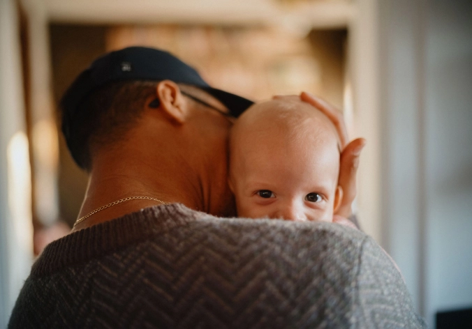 A man holds a baby close to him while the baby peeks over his shoulder.