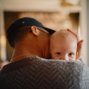 A man holds a baby close to him while the baby peeks over his shoulder.