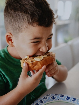 A smiling boy sitting at a table eating a piece of bread with peanut butter and toppings.