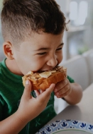 A smiling boy sitting at a table eating a piece of bread with peanut butter and toppings.