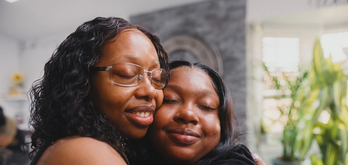 A Black woman with glasses hugs her daughter in their new home.