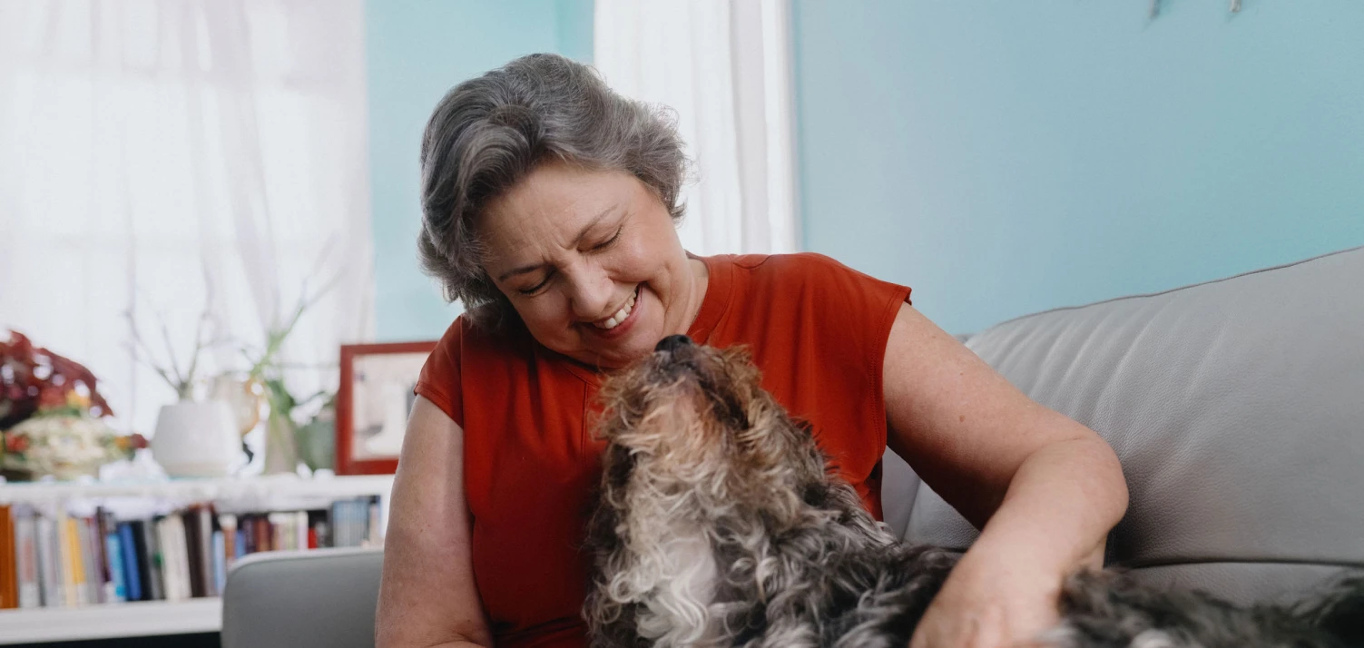 A woman with gray hair pets her dog while sitting on the couch in her living room.