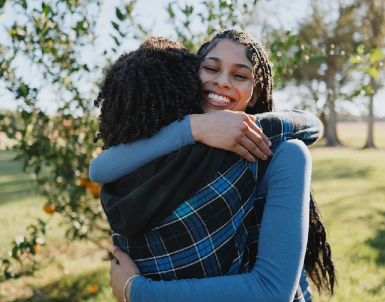 A multicultural young woman with long braided hair hugs a friend at a meetup at an orchard.