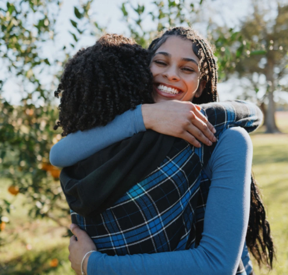 A multicultural young woman with long braided hair hugs a friend at a meetup at an orchard.