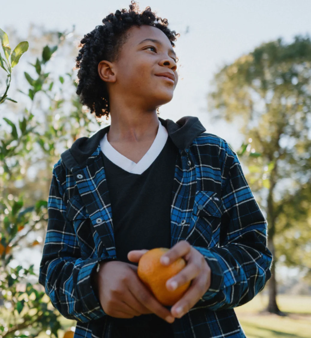 Un niño con una camisa a cuadros azul y negra que visita un huerto sostiene una naranja que ha recogido.