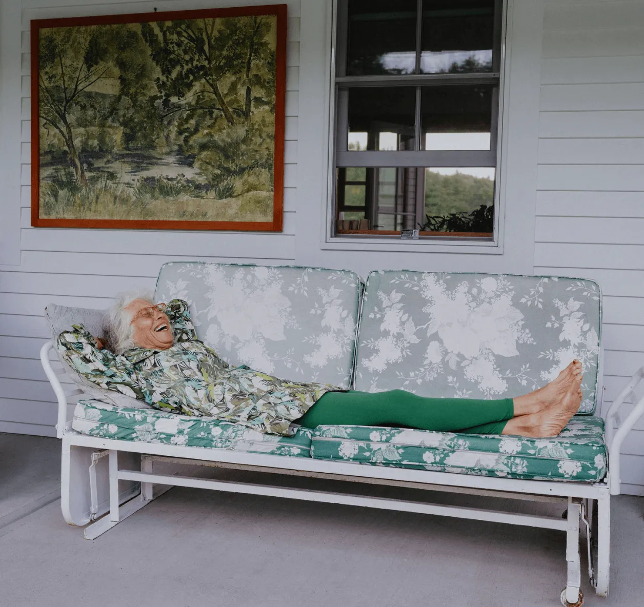 A woman with white hair relaxes on a porch swing in front of a white house that she purchased with Rocket Mortgage.