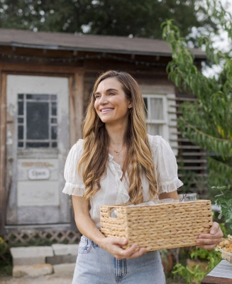 A white woman stands in front of her small business, holding a basket of handmade goods.