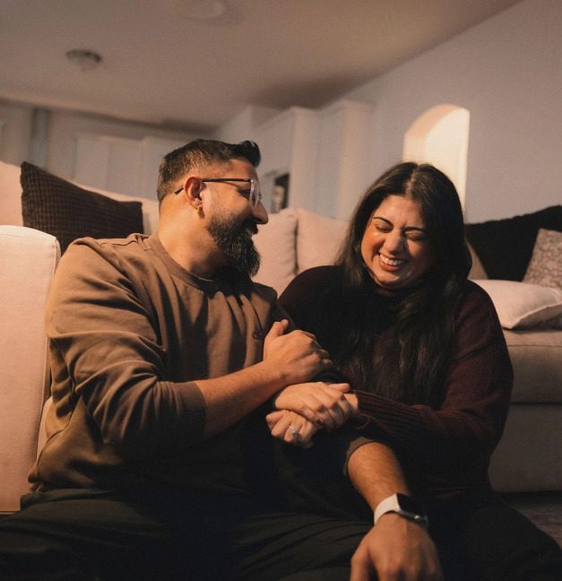 Una pareja hispana se sienta en el suelo de su nuevo hogar, tomados de la mano y riendo.