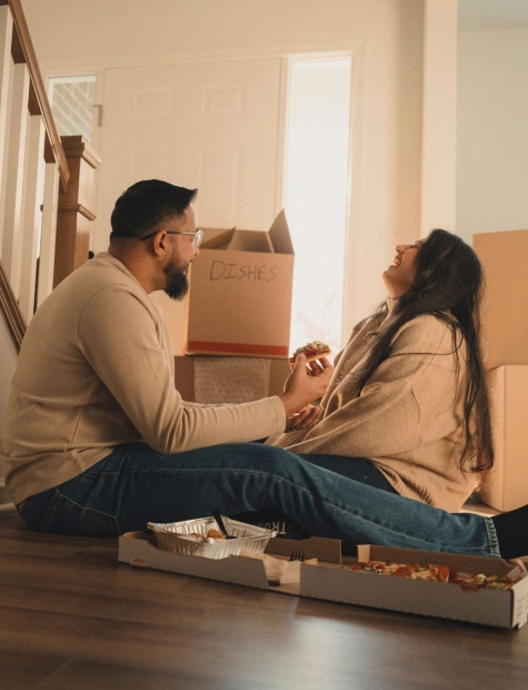 Una pareja hispana se sienta en el suelo de su nuevo hogar rodeada de cajas de mudanza, comiendo pizza y riendo.