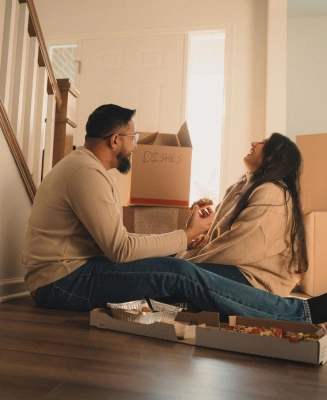 Una pareja hispana se sienta en el suelo de su nuevo hogar rodeada de cajas de mudanza, comiendo pizza y riendo.