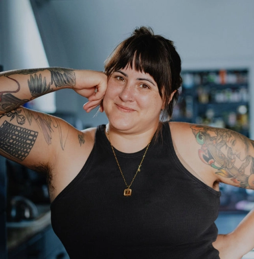 A young white woman with tattoos on her bare arms wearing a black tank top poses confidently.