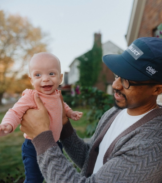 A young Black dad in a cardigan wearing a baseball cap holds his 3 month old smiling baby.