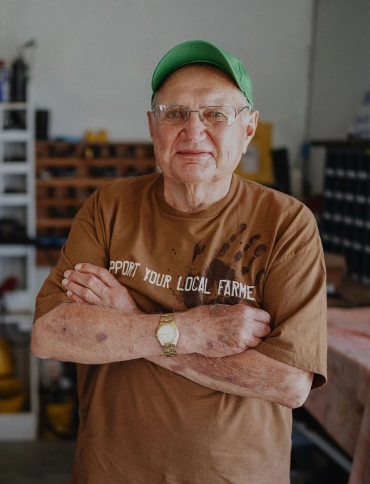 An elderly white man in a green baseball cap wears a brown t-shirt that says support your local farmer.