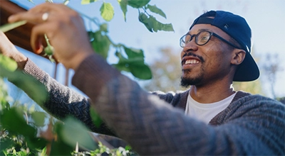 A Black man wearing glasses and a backwards baseball cap examines some greenery around his home.