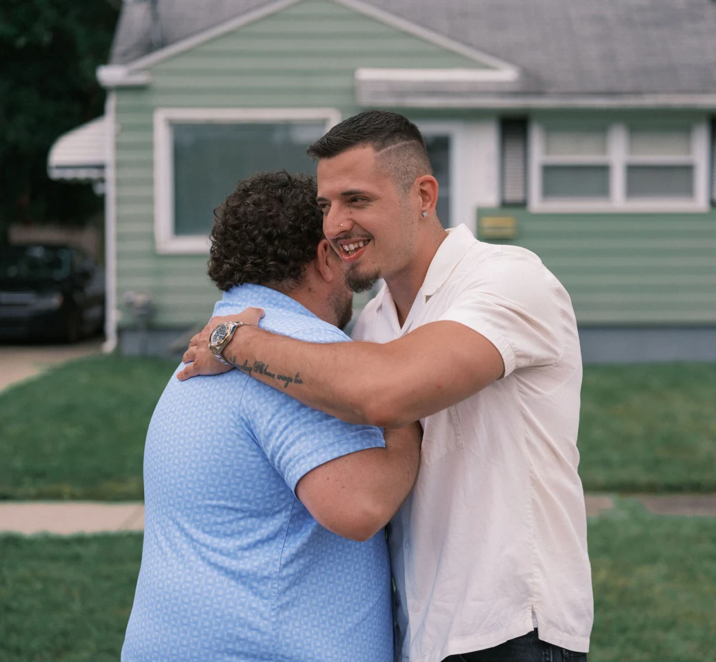 A man in his 30s with a goatee, wearing a white polo shirt, hugs a neighbor wearing a blue shirt in front of a home with light green siding.