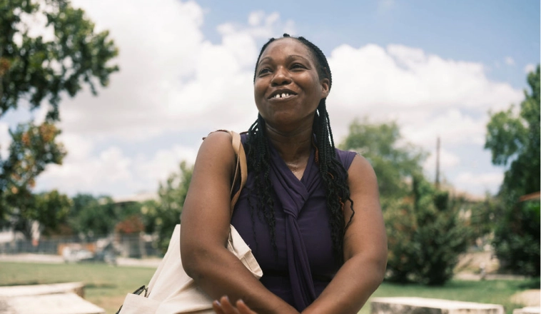 A Black woman with long braided hair and a tote bag over her shoulder looks engaged in conversation outdoors.