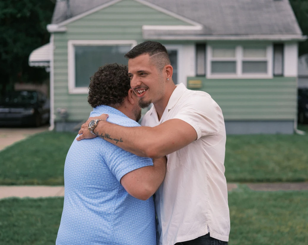 Un hombre de unos 30 años con perilla y vestido con un polo blanco, abraza a un vecino que viste una camisa azul frente a una casa con revestimiento verde claro.