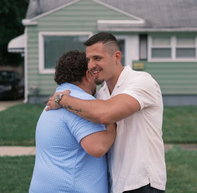 A man in his 30s with a goatee, wearing a white polo shirt, hugs a neighbor wearing a blue shirt in front of a home with light green siding.