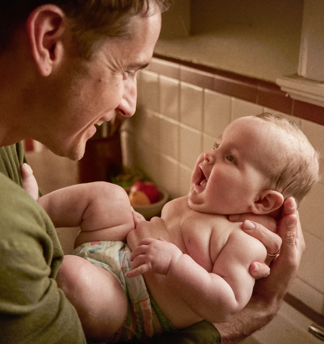 A white man holds a baby in a diaper in his arms while they both smile at each other.