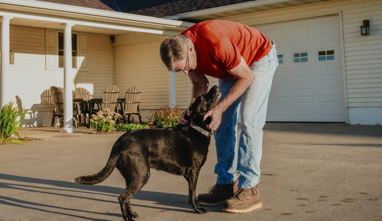 A mature white man in jeans, a red shirt and work boots leans over to pet his dog in the driveway of his home.