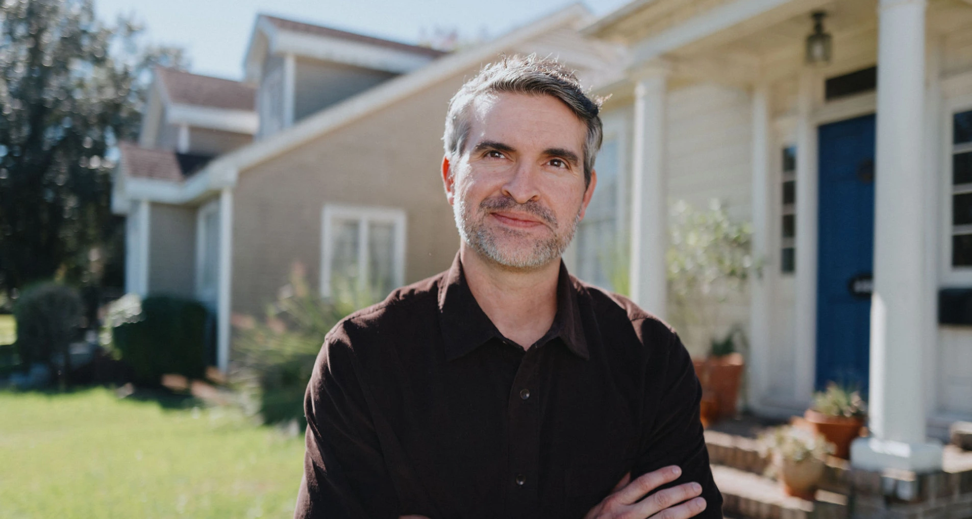 A middle-aged white man in a dark brown shirt stands with his arms crossed in front of his home.