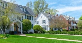 Three neighborhood homes in a row: a white colonial with green shutters and a large green lawn, another white home, and a brick home. 