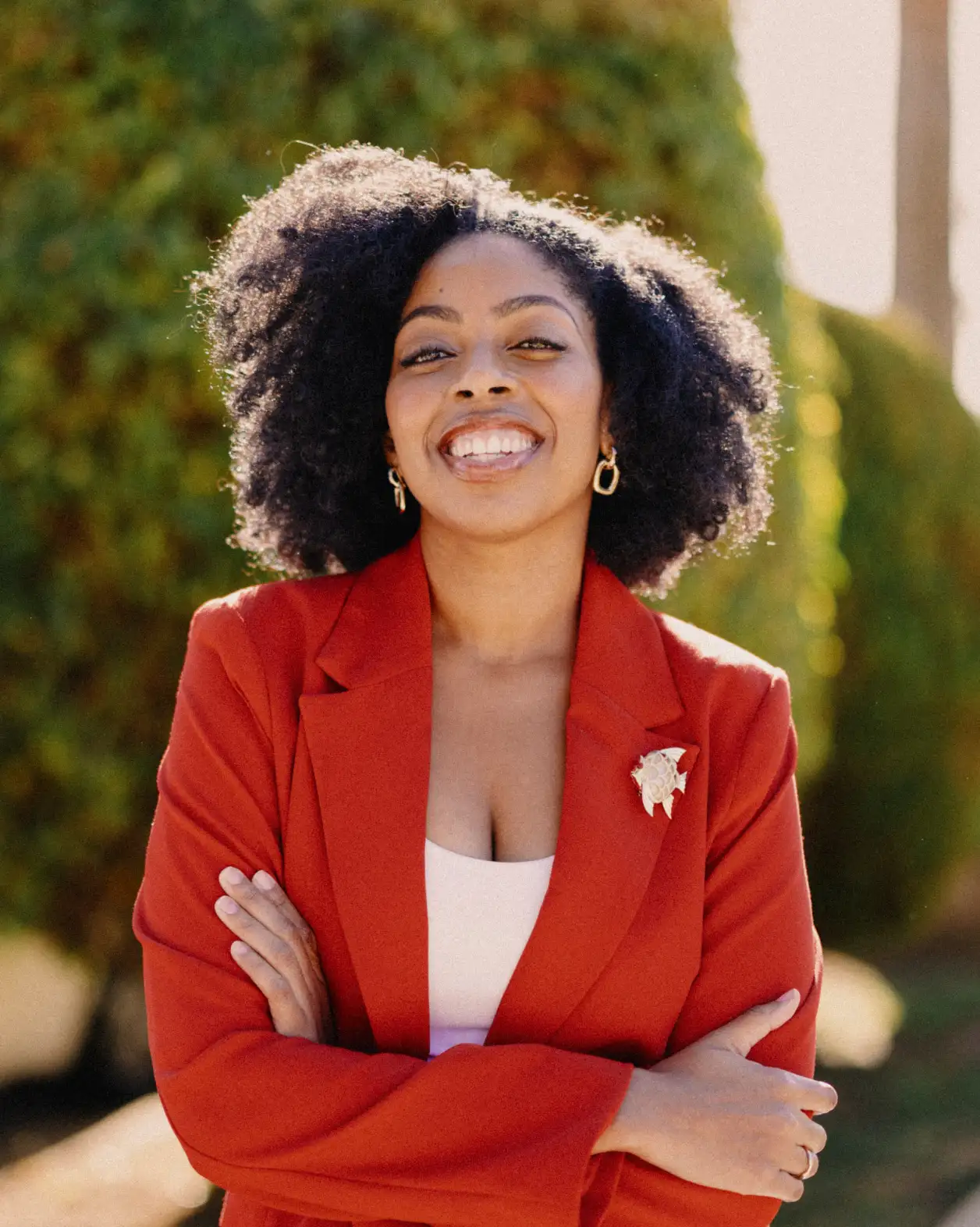 A woman with black curly hair stands confidently and smiles at the camera while crossing her arms. She is wearing a red blazer with a gold broach. 