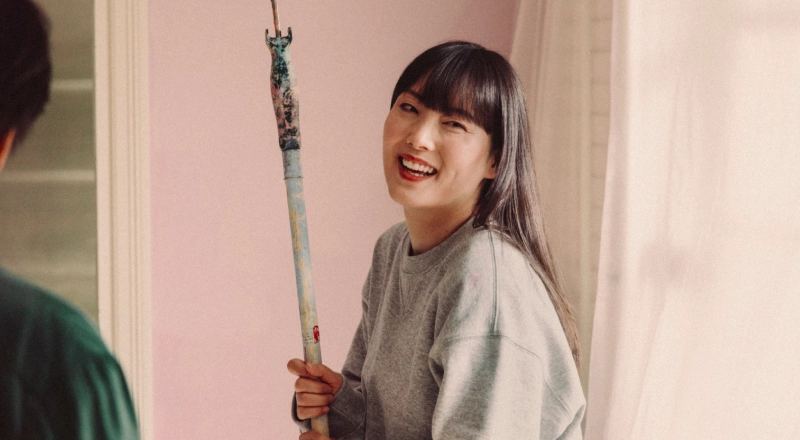 A woman with dark hair holds a paint roller while painting a pink room.