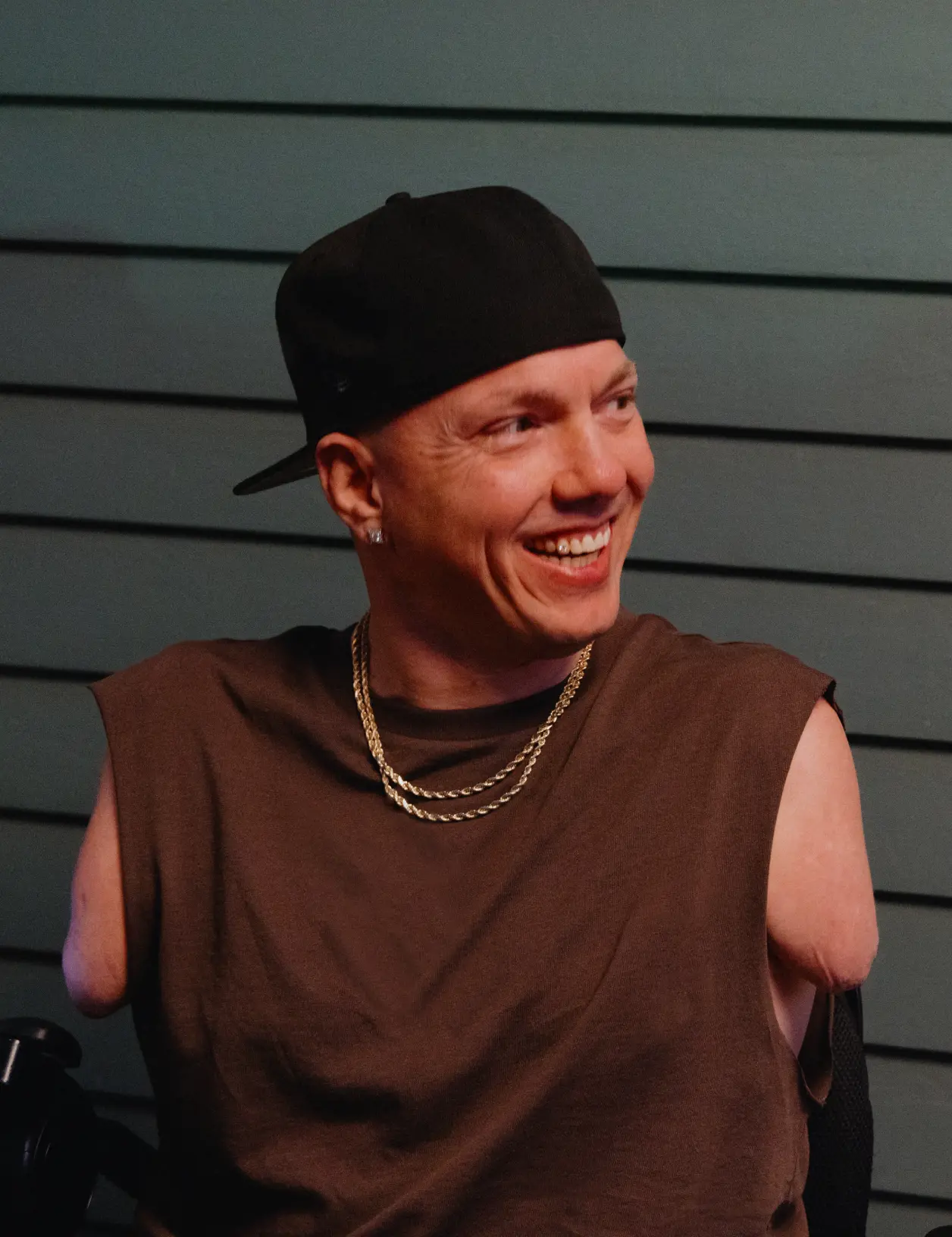 A white man with no arms wearing a backwards ball cap and gold chains smiles while looking to his left.