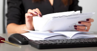 A woman holding a stack of envelopes and papers over a computer keyboard and mouse.