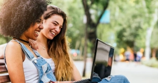 A young Black woman in denim overalls and her friend, a young white woman with long hair, smile while looking at a laptop together on a park bench.