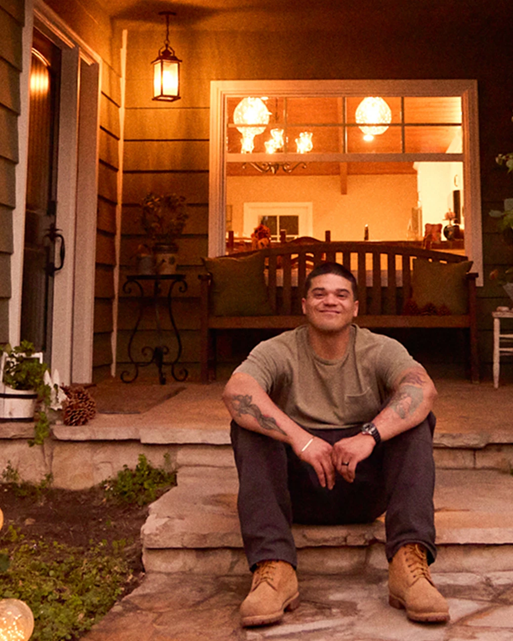 A Hispanic man wearing work boots, brown pants and a tan t-shirt smiles proudly while sitting on the front steps of a stone porch, with a lighted picture window in the background.