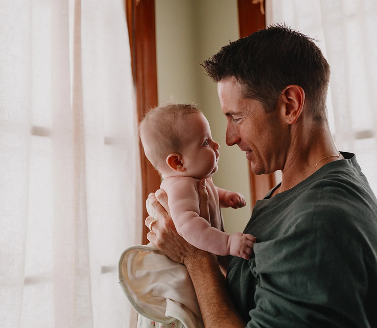 A clean-shaven white man with short dark hair holds a baby up close to his face and stares lovingly into the baby’s eyes.