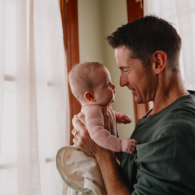 A clean-shaven white man with short dark hair holds a baby up close to his face and stares lovingly into the baby’s eyes.