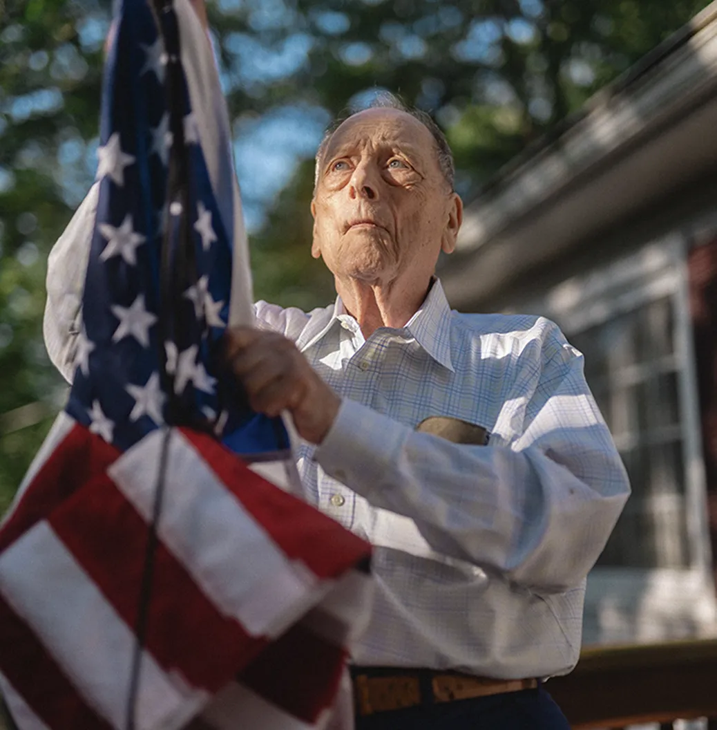 Robert B., an elderly veteran wearing a white shirt holds the American flag.