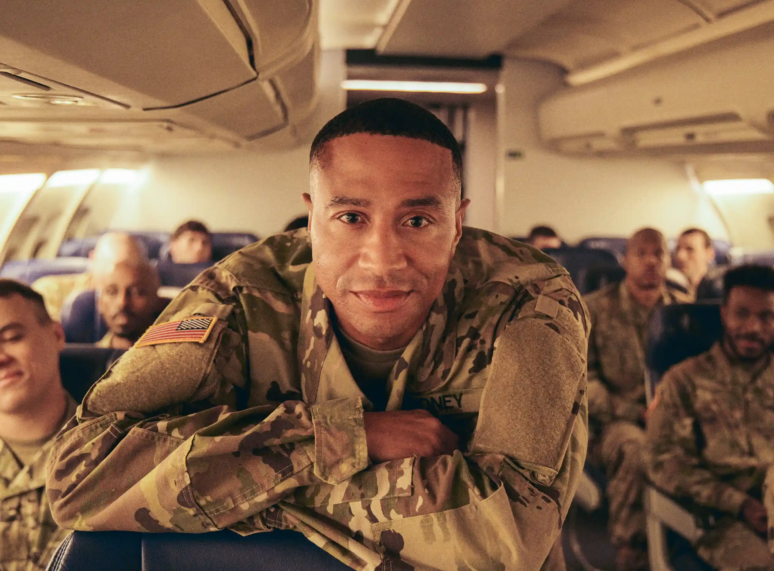 A Black soldier in a military uniform leans over the top of an airline seat on a flight with other soldiers.