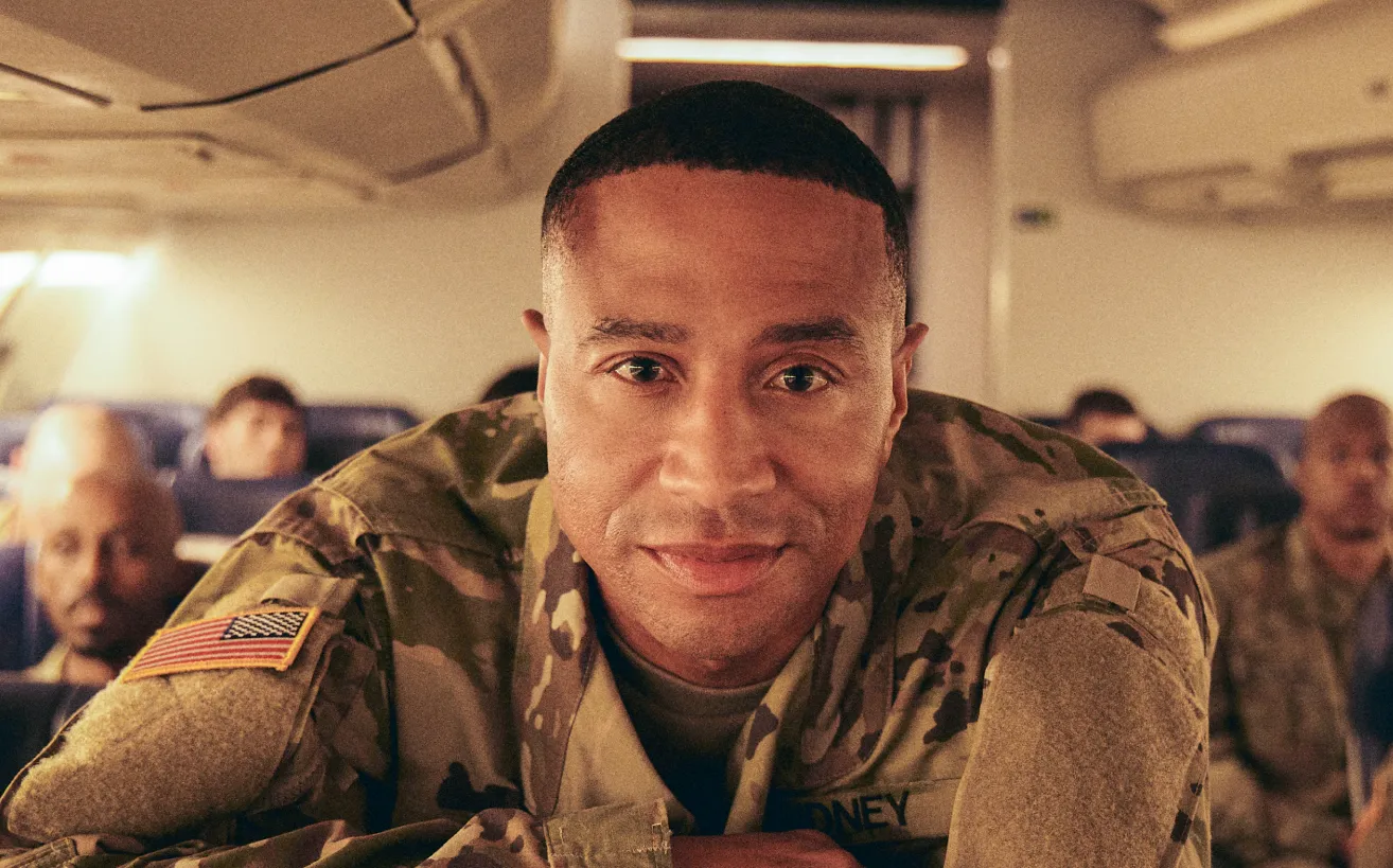 A Black soldier in a military uniform leans over the top of an airline seat on a flight with other soldiers.